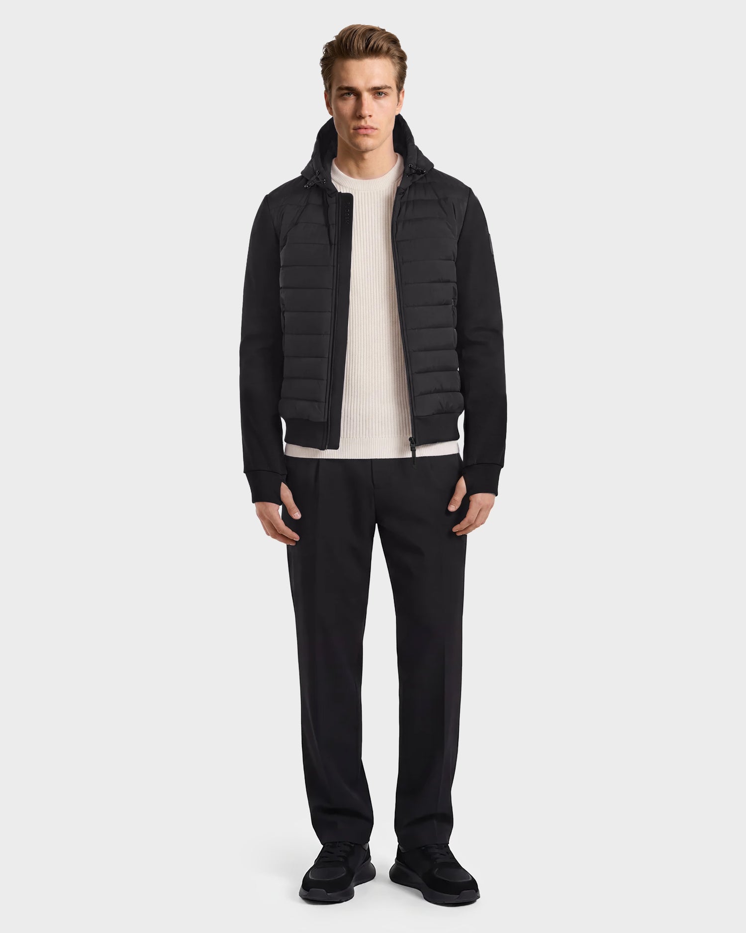 A person stands facing forward against a plain light background, wearing the Rudsak DEE MAT - 6124028 Black hybrid down jacket over a white ribbed sweater, black pants, and black shoes.