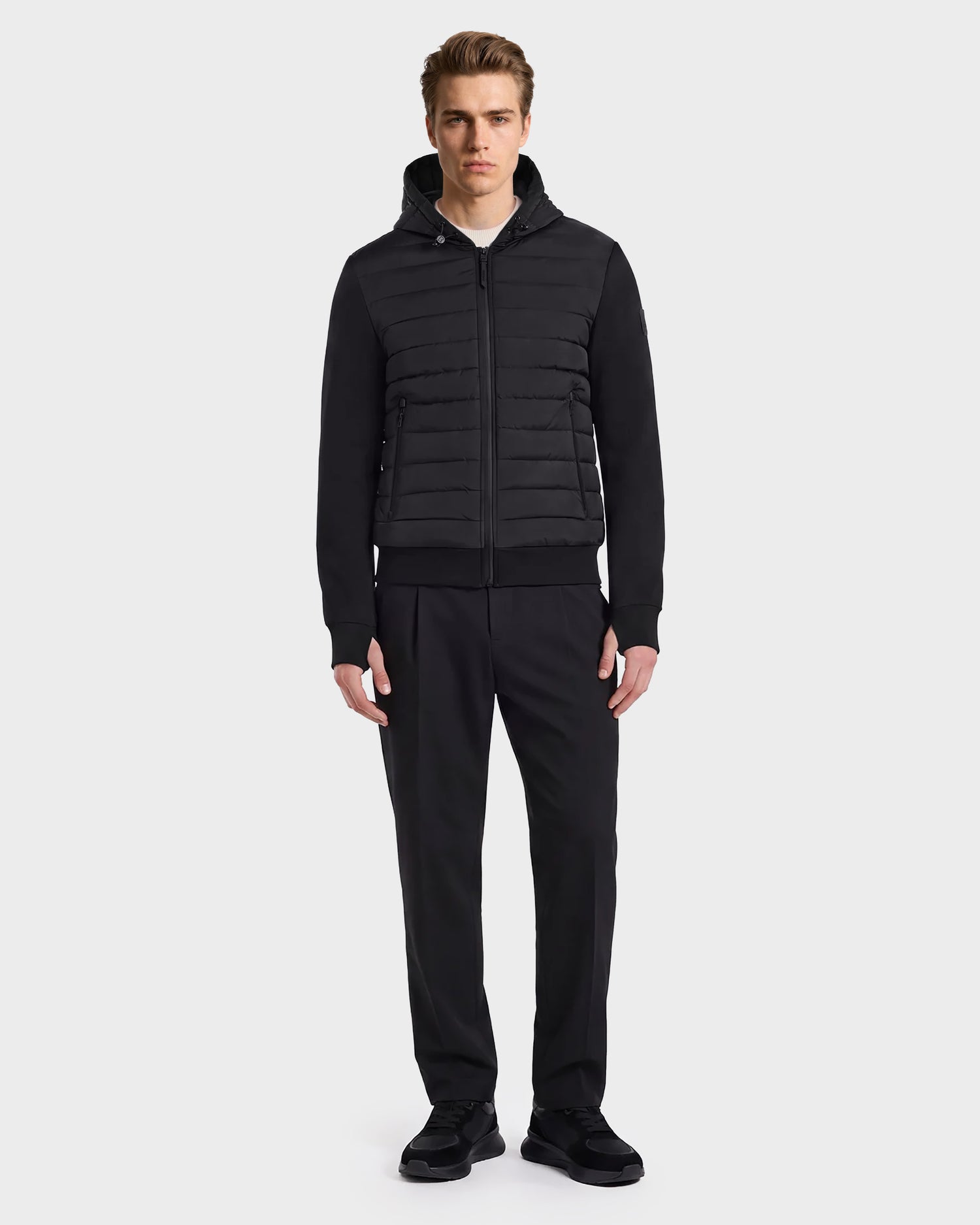 A man stands facing forward, wearing the Rudsak DEE MAT - 6124028 Black hybrid down jacket with horizontal quilting, paired with black pants and black shoes, against a plain light gray background.