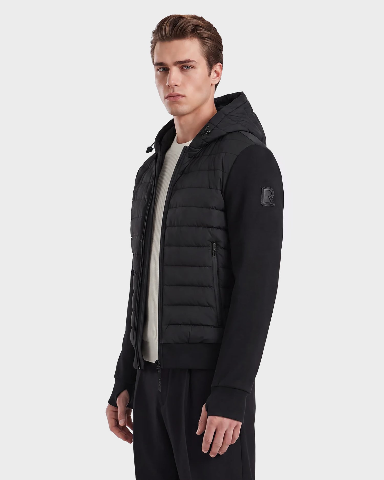 A man wearing the Rudsak DEE MAT - 6124028 black hybrid down jacket, featuring quilted front panels, black sleeves, and a front zipper, stands facing slightly to the side against a plain light gray background.
