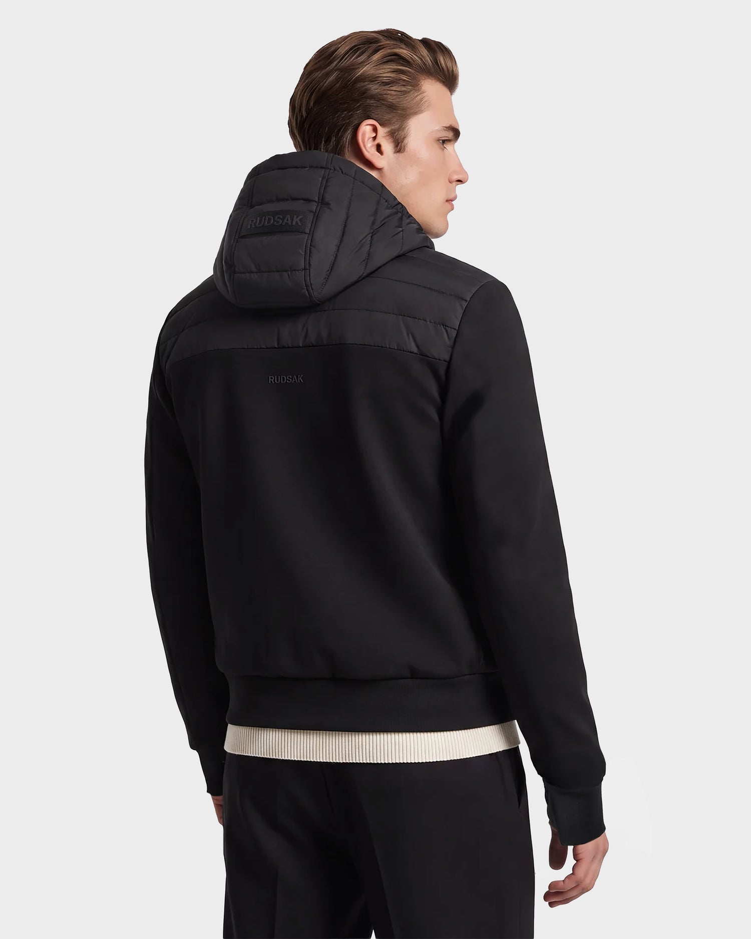 A man with brown hair models the Rudsak DEE MAT - 6124028 Black men's lightweight down jacket, which features a quilted hood and RUDSAK branding on the back. He stands against a plain light background, facing away from the camera.