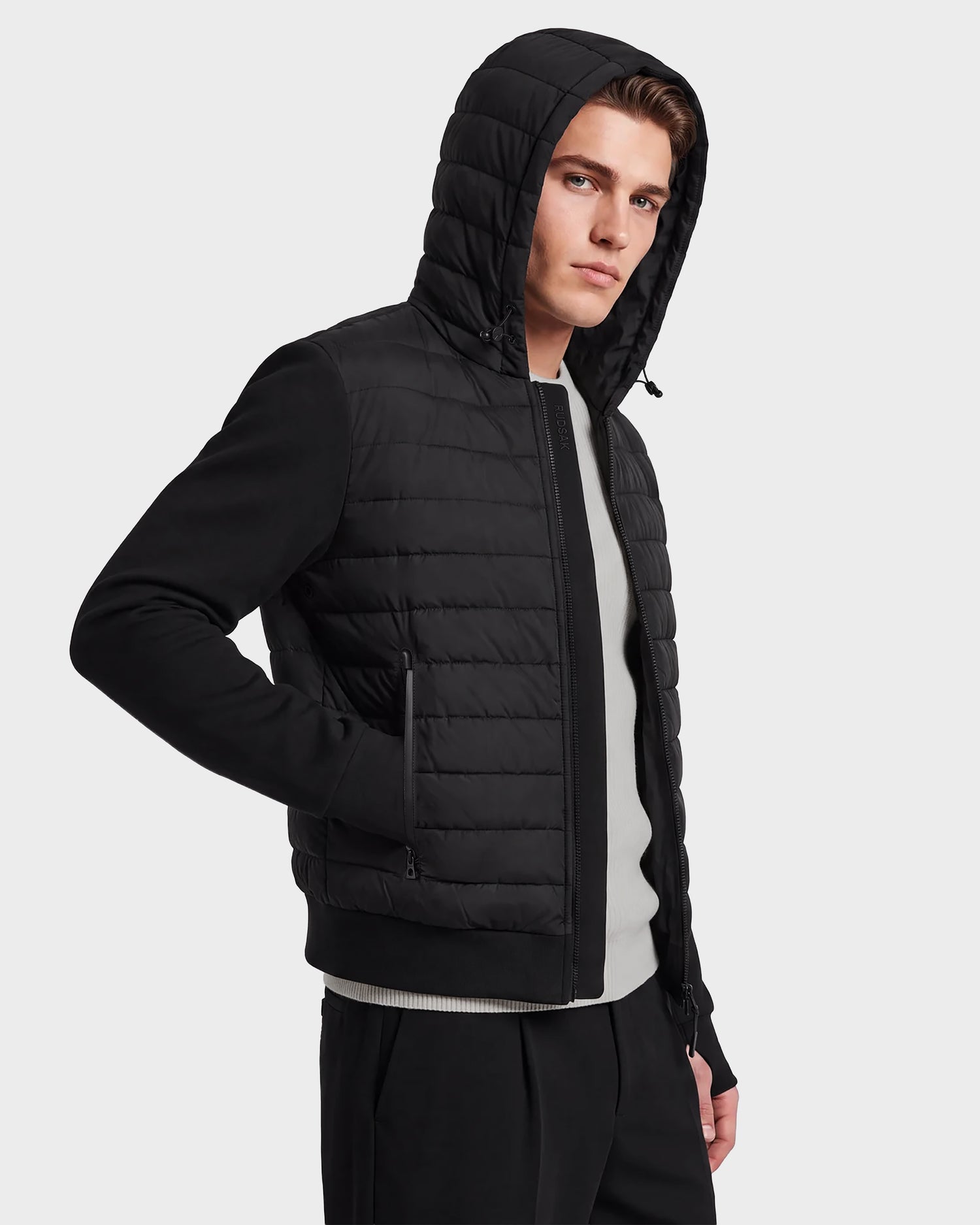 A man stands against a plain background with his right hand in his pocket, looking slightly to the left while wearing the Rudsak DEE MAT - 6124028 Black men's lightweight down hooded puffer jacket over a light gray shirt.
