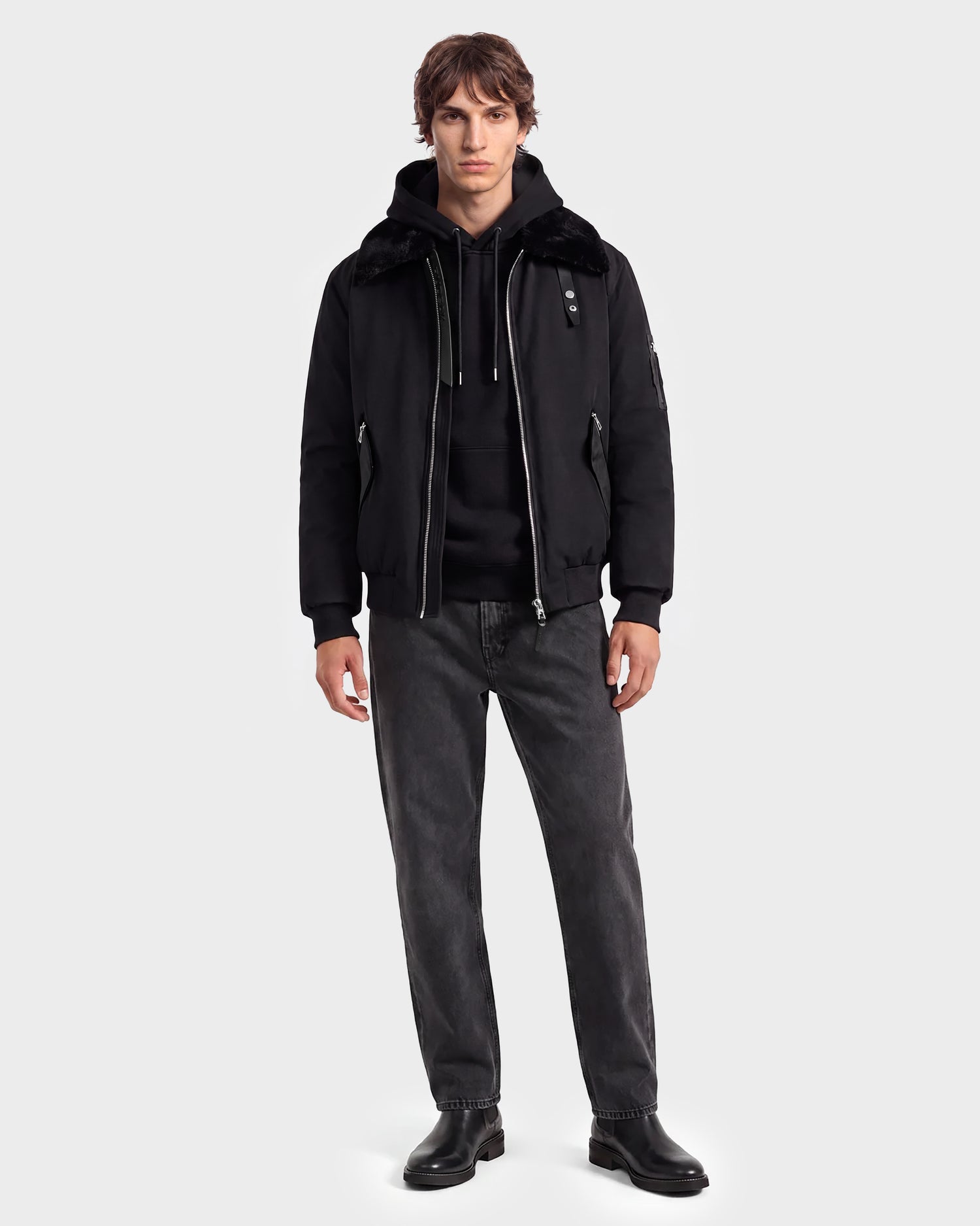 A person stands facing forward against a plain light gray background, effortlessly showcasing modern outerwear by wearing the Rudsak DYLAN - 6125593 Black men’s aviator bomber jacket with a removable faux fur collar over a black hoodie, paired with dark gray jeans and black boots.