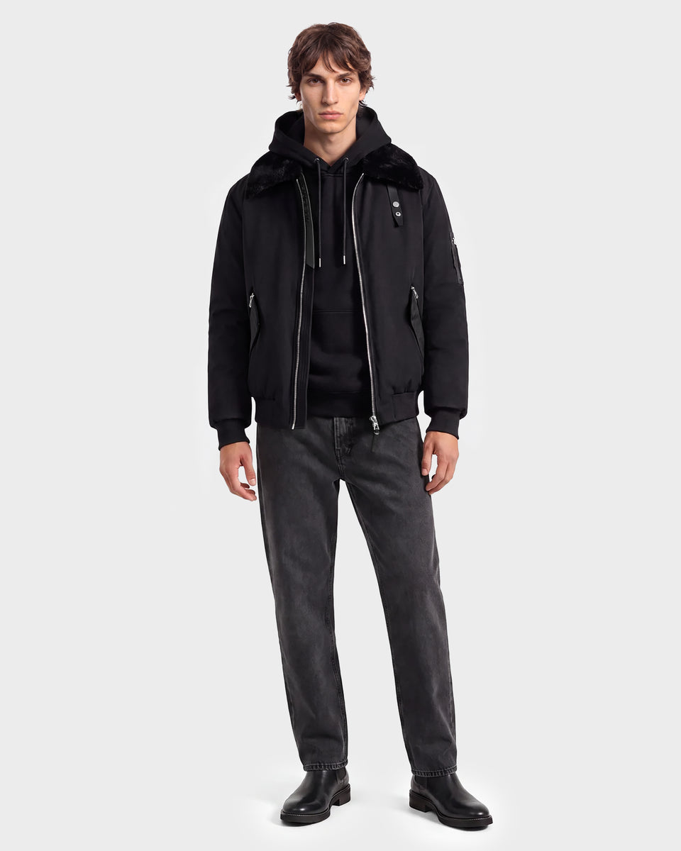 A person stands facing forward against a plain light gray background, effortlessly showcasing modern outerwear by wearing the Rudsak DYLAN - 6125593 Black men’s aviator bomber jacket with a removable faux fur collar over a black hoodie, paired with dark gray jeans and black boots.