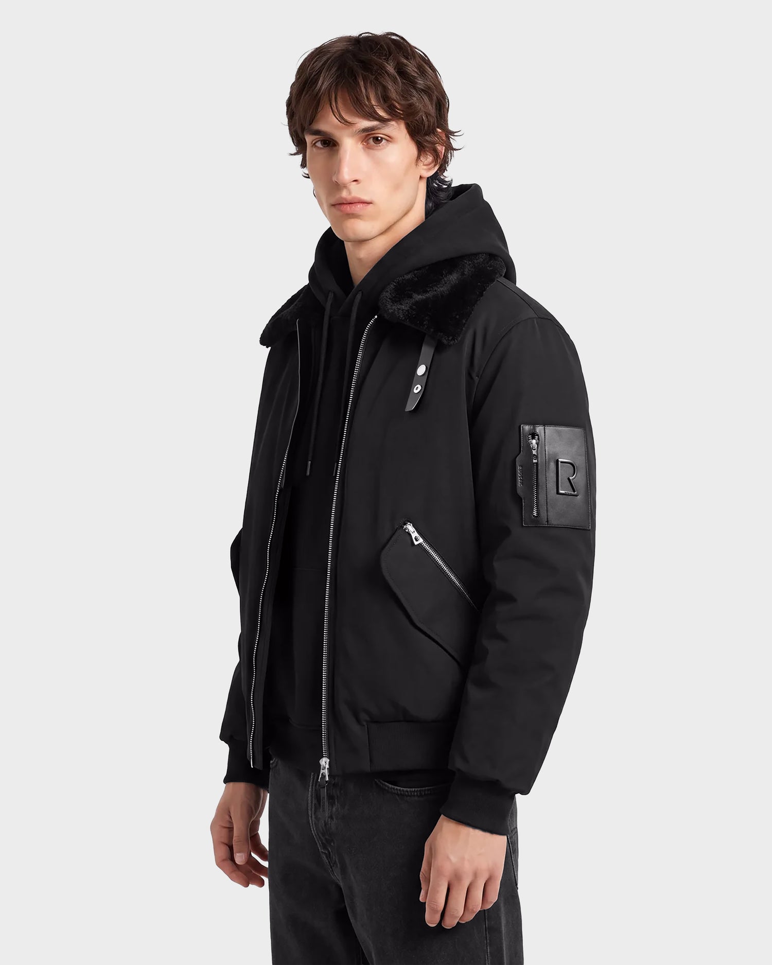 A person stands facing slightly left against a plain light background, wearing the Rudsak DYLAN - 6125593 Black men’s aviator bomber jacket with a modern outerwear design, featuring a removable faux fur collar, zippered pockets, and an R patch on the sleeve.