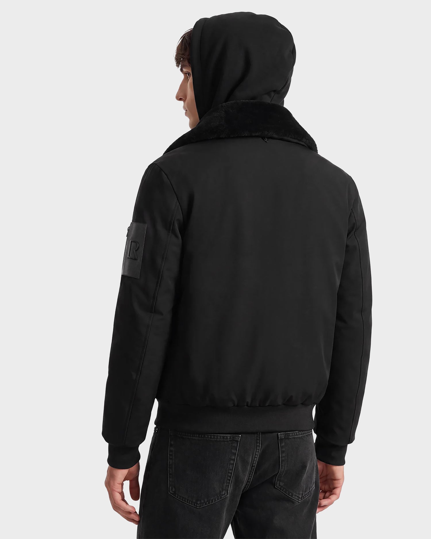 A person is seen from behind, looking to the left against a plain light background, wearing the Rudsak DYLAN - 6125593 Black men's aviator bomber jacket with a removable faux fur collar and black jeans.
