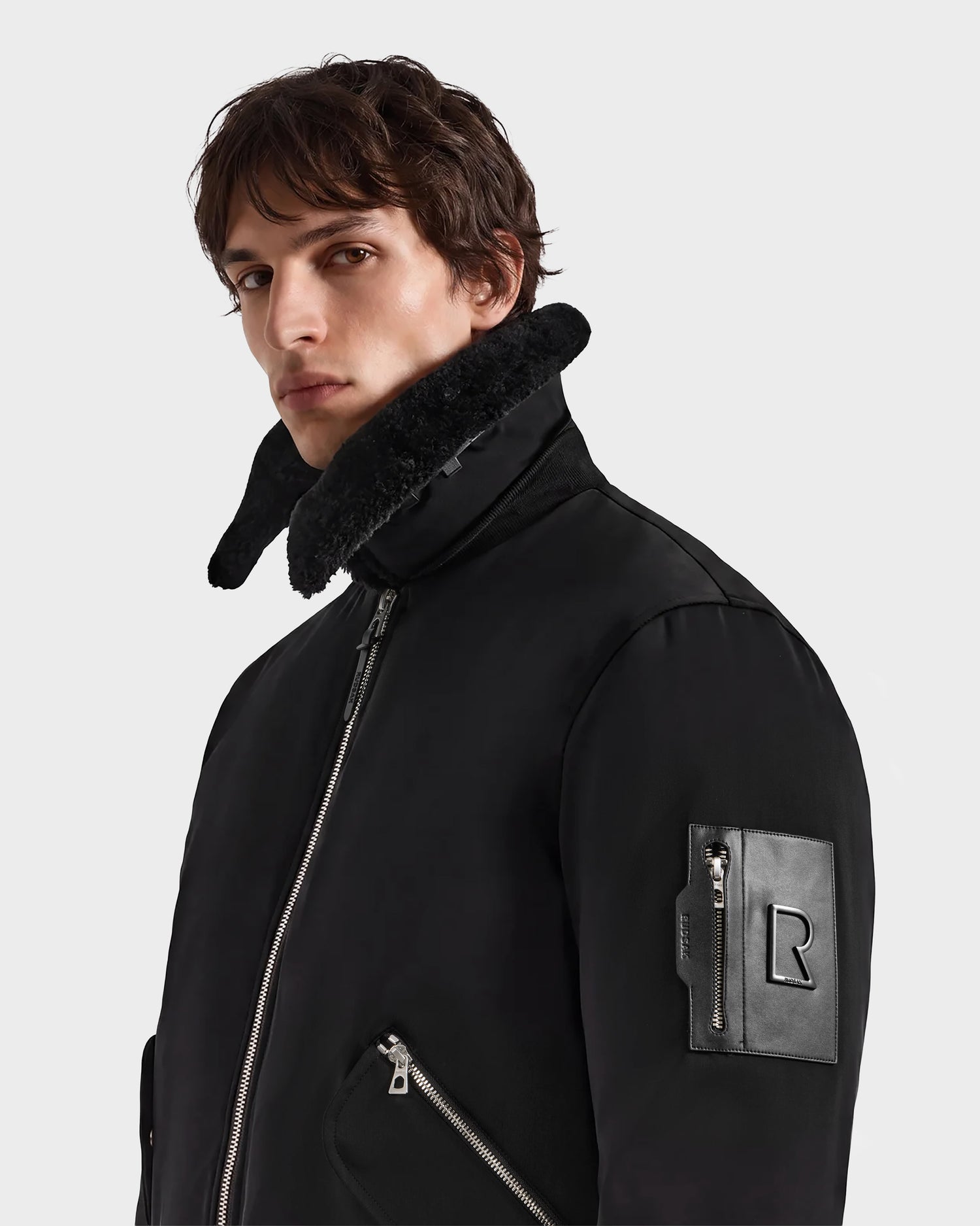 A man wearing the DYLAN - 6125593 Black men’s aviator bomber jacket by Rudsak, featuring a high shearling collar and zippered pockets, stands against a plain light background, looking toward the camera. This modern outerwear piece includes a removable faux fur collar for versatile styling.
