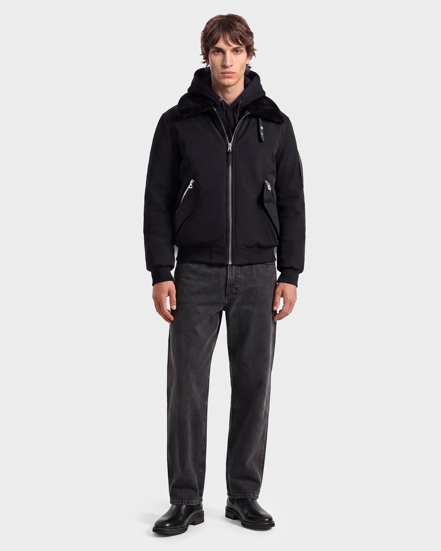 A man stands facing forward, wearing the Rudsak DYLAN - 6125593 Black modern outerwear zip-up jacket with a removable faux fur collar over a black hoodie, paired with dark gray jeans and black shoes against a plain light background.