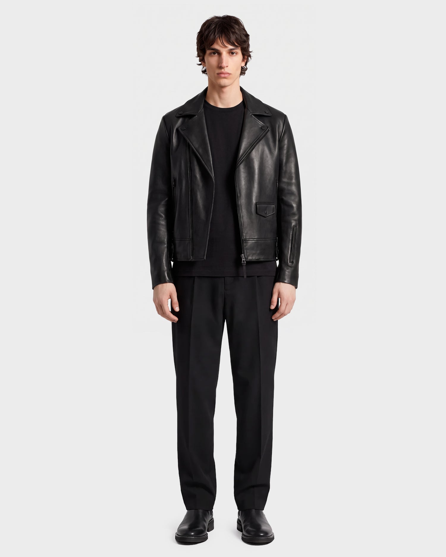 A person stands facing forward against a plain light background, wearing RUDSAK’s DAREG - 6122004 BLACK lamb leather biker jacket, paired with a black shirt, black pants, and black shoes—a true wardrobe essential.