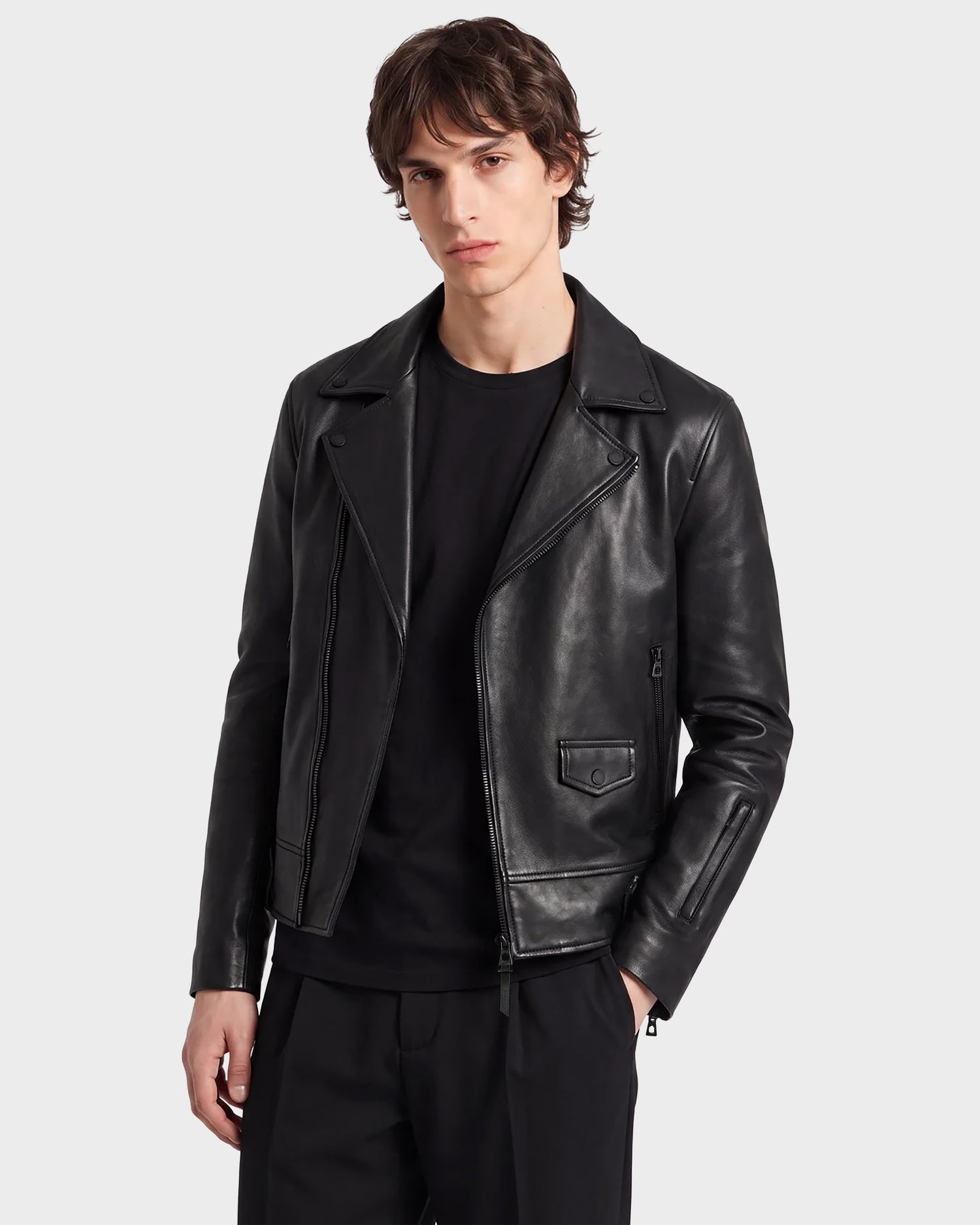 A person with medium-length brown hair wears the RUDSAK DAREG - 6122004 BLACK jacket—an undeniable wardrobe essential—crafted from supple lamb leather, layered over a black shirt and black pants, standing against a plain light gray background.