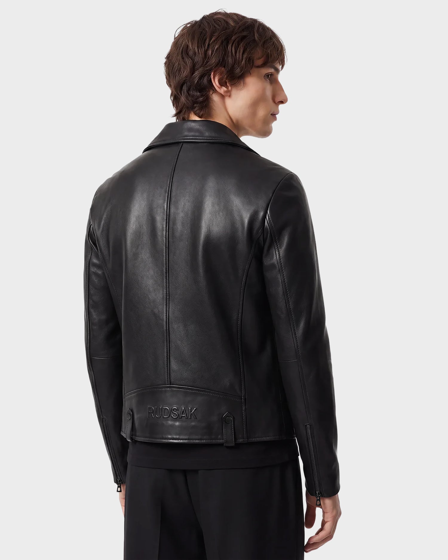 A person with medium-length brown hair wears the RUDSAK DAREG - 6122004 BLACK lamb leather biker jacket, featuring RUDSAK embossed on the lower back—a true wardrobe essential—standing against a plain light background and facing away from the camera.