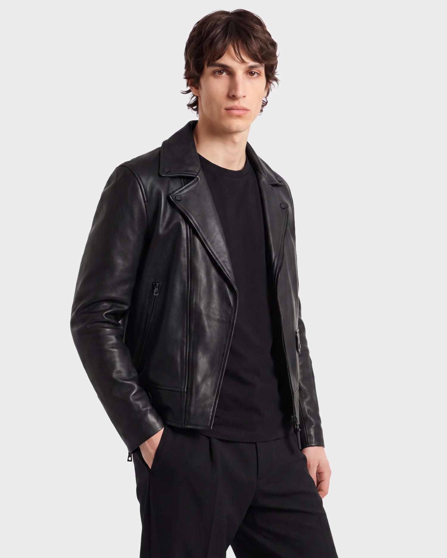 A person stands facing forward against a plain light background, wearing RUDSAK’s DAREG - 6122004 BLACK lamb leather biker jacket—a true wardrobe essential—styled over a black shirt and black pants, with one hand in their pocket.