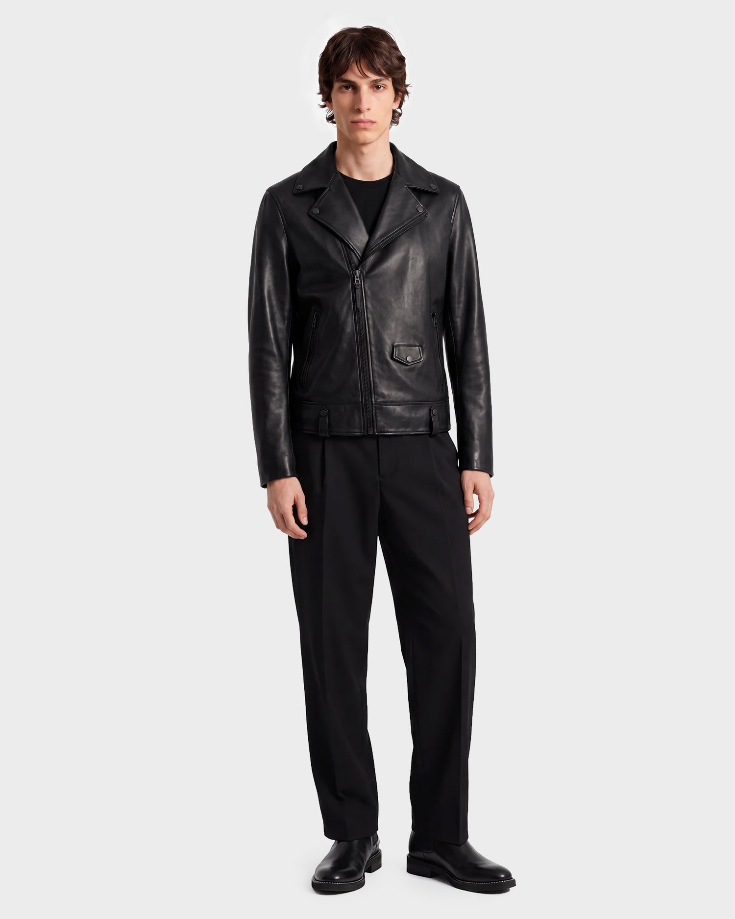 A person stands wearing the RUDSAK DAREG - 6122004 BLACK lamb leather biker jacket, paired with black shirt, pants, and shoes against a plain light background—a true wardrobe essential.