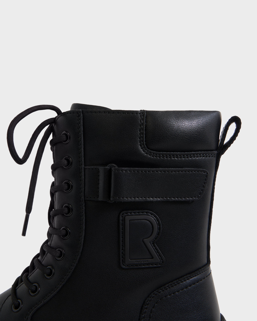 A close-up of the DEREK - 6226506 Black combat boot by Rudsak features black laces, a hook-and-loop strap, and a raised R logo, highlighting sleek winter style against a plain white background.