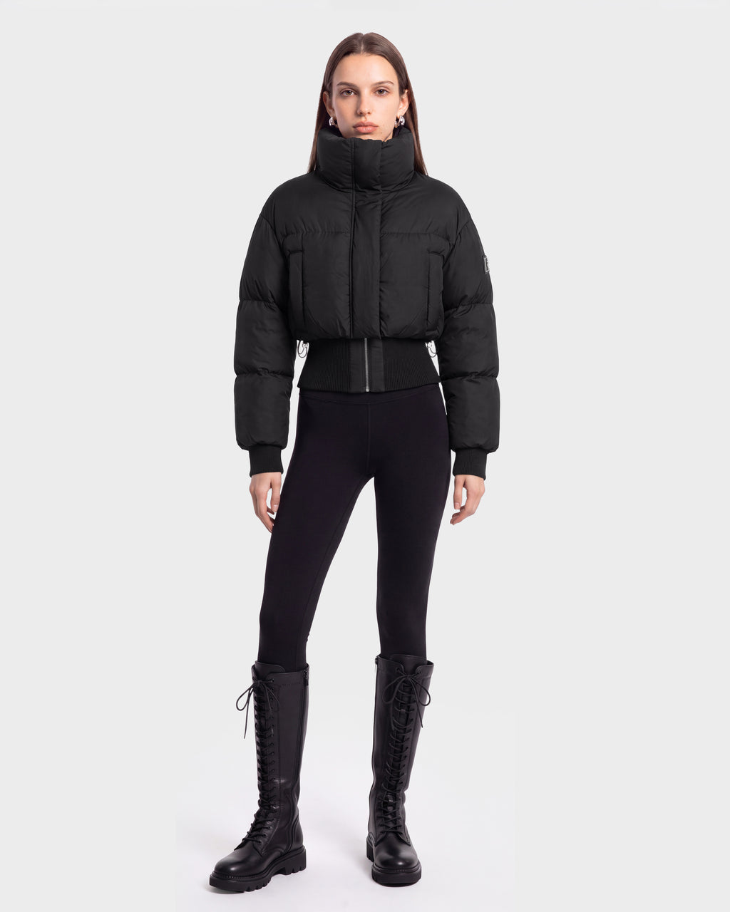 A person stands facing forward, wearing the Rudsak DONYA - 8126516 Black puffer jacket, styled with black leggings and tall black lace-up boots against a plain light gray background. This look is available for pre-order and is expected to ship by September 15th.