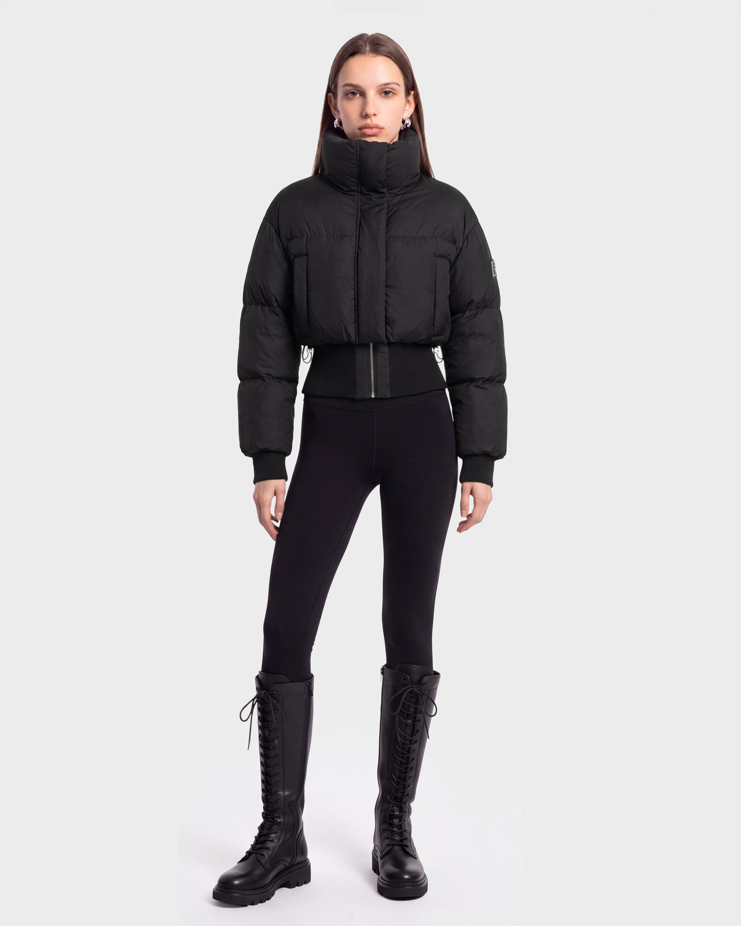 A person stands facing forward, wearing the Rudsak DONYA - 8126516 Black puffer jacket styled with black leggings and black knee-high lace-up boots against a plain light gray background. This stylish Rudsak outfit is available for pre-order now and is expected to ship by September 15th.