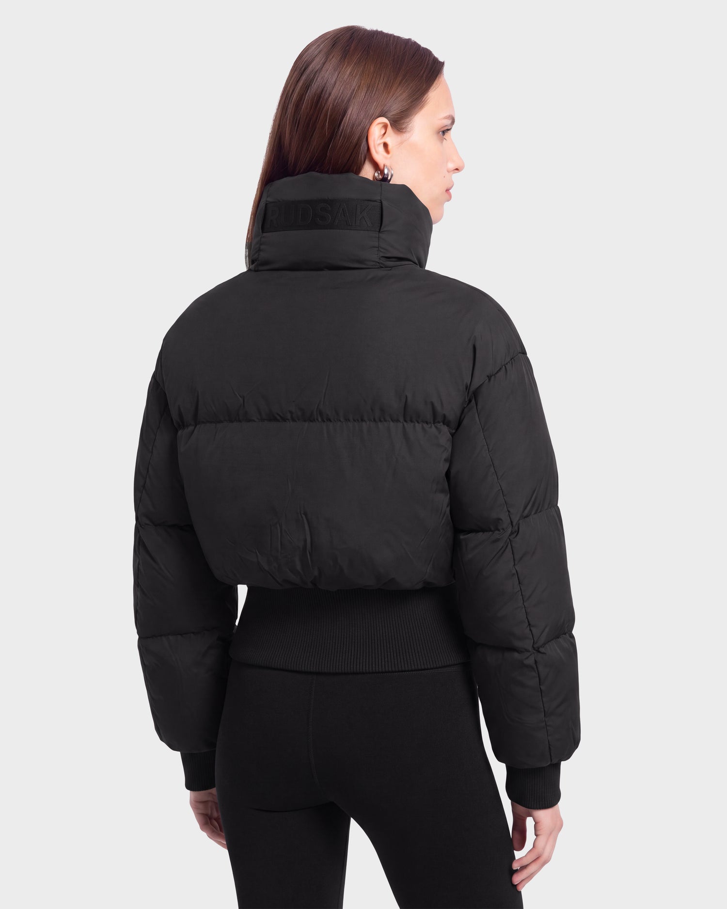 A woman with straight brown hair, seen from behind against a plain light background, wears the Rudsak DONYA - 8126516 Black puffer jacket paired with black leggings. This item is available for pre-order and is expected to ship by September 15th.