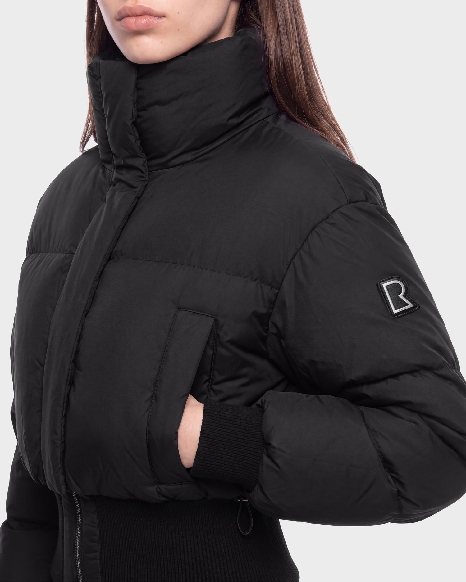 A person wearing the Rudsak DONYA - 8126516 Black puffer jacket, featuring a high collar, ribbed cuffs, and a signature B logo on the sleeve, stands against a plain light background. This item is available for pre-order and is expected to ship by September 15th.