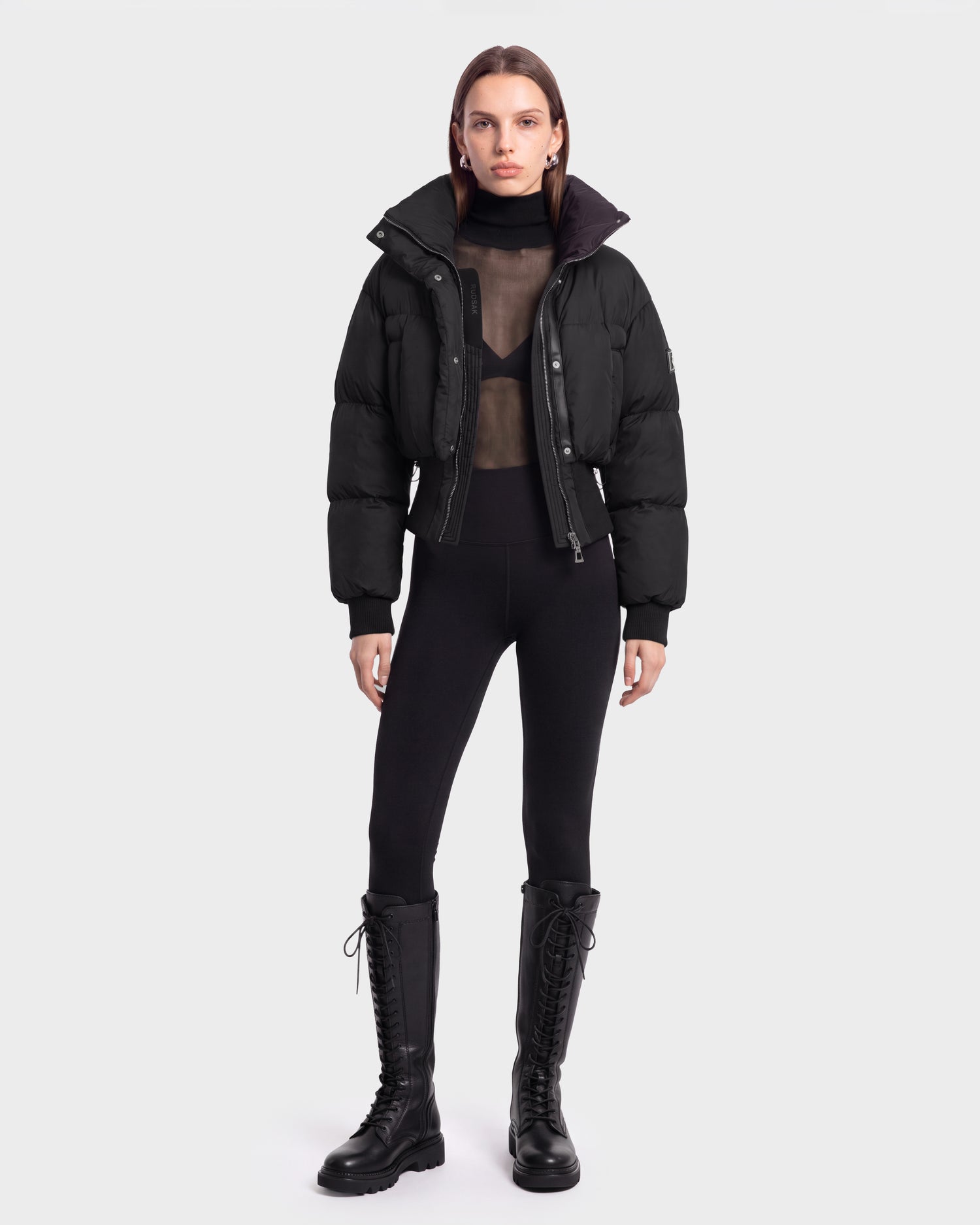 A person wearing the Rudsak DONYA - 8126516 Black puffer jacket, paired with a sheer black top, black leggings, and black lace-up boots, stands facing forward against a plain light background. This stylish ensemble is available for pre-order now and is expected to ship by September 15th.