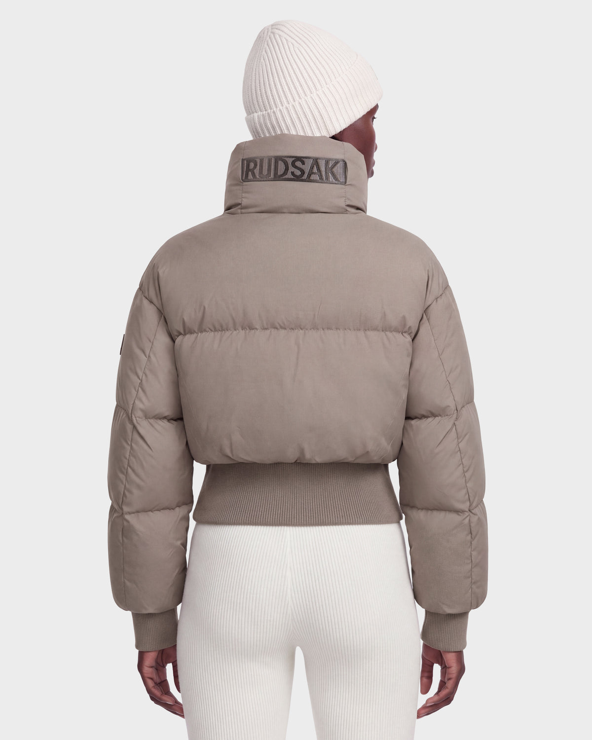 A woman is shown from the back against a plain light background, wearing Rudsak’s DONYA - 8126516 Cement women’s down puffer jacket featuring RUDSAK branding on the high collar, paired with white ribbed pants and a white knit beanie. This RDS-certified winter jacket combines warmth and style for comfortable cold-weather wear.