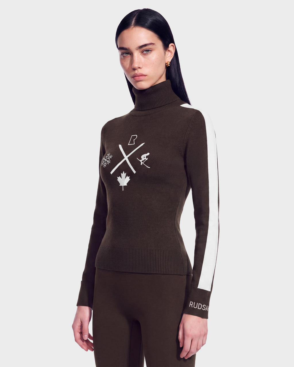 A woman with long dark hair wears the Rudsak ELIZABETH - 8524623 Espresso women’s ski turtleneck sweater, featuring white symbols and a white stripe down the sleeve, paired with matching brown pants, standing against a plain background—an effortless addition to any winter wardrobe.