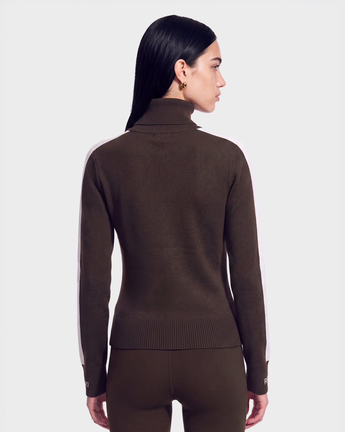 A woman with straight black hair models the Rudsak ELIZABETH - 8524623 Espresso après-ski turtleneck sweater featuring white stripes down the sleeves, paired with matching pants. She is shown from the back against a plain light background.