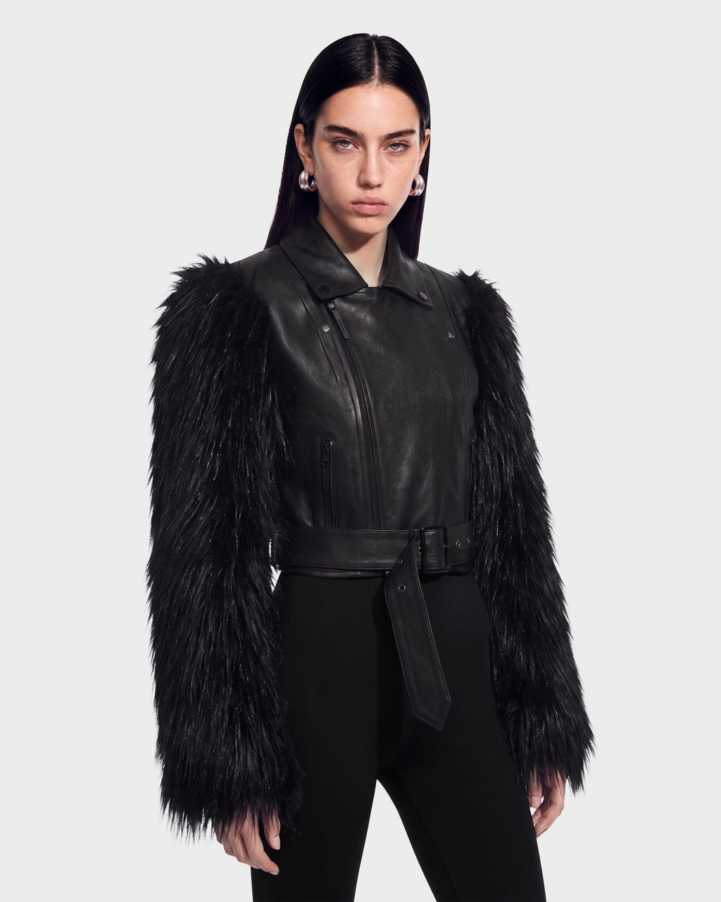 A woman stands against a plain light gray background wearing the Rudsak EMELDA - 8126991 Black leather moto jacket featuring long, luxe faux fur sleeves, paired with black pants and silver hoop earrings.