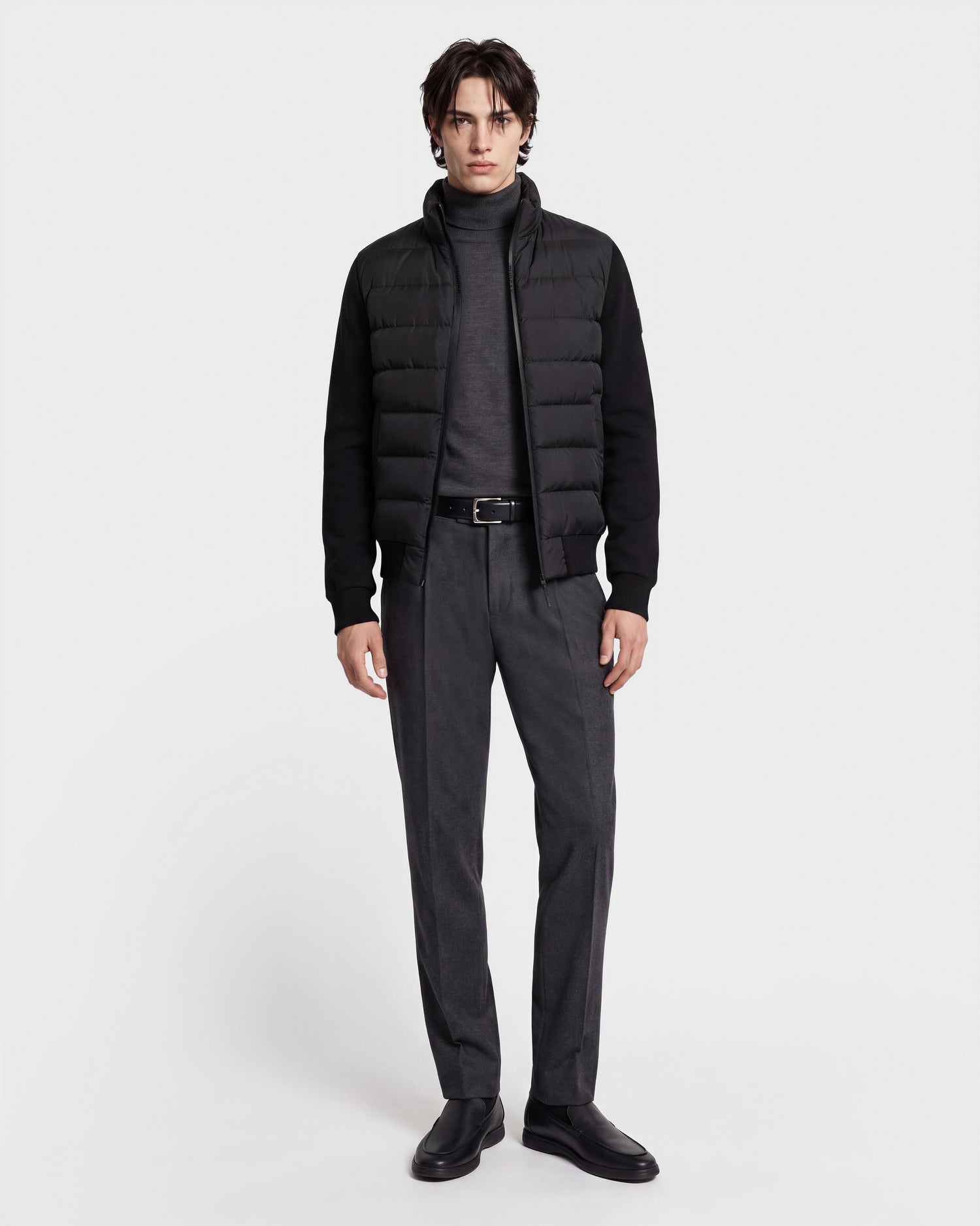 A person stands facing forward against a plain white background, wearing the Rudsak EUGENE - 6124721 Black performance luxury jacket layered over a gray turtleneck sweater, paired with gray dress pants, a black belt, and black shoes.