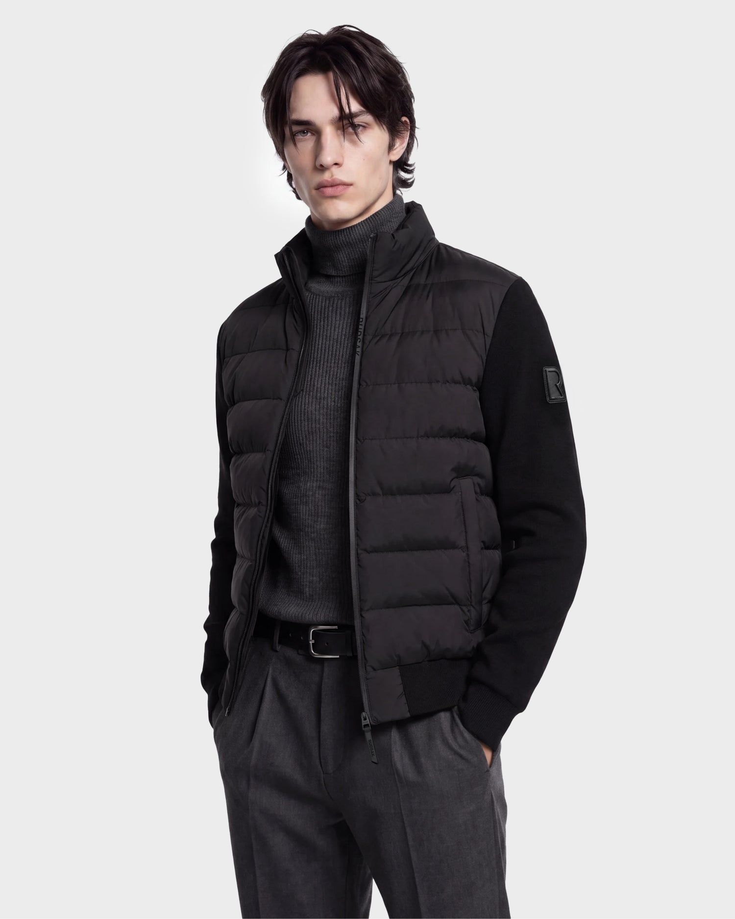 A man stands with his hands in his pockets against a plain light background, wearing the Rudsak EUGENE - 6124721 Black lightweight mixed-media jacket layered over a charcoal turtleneck and gray pants.