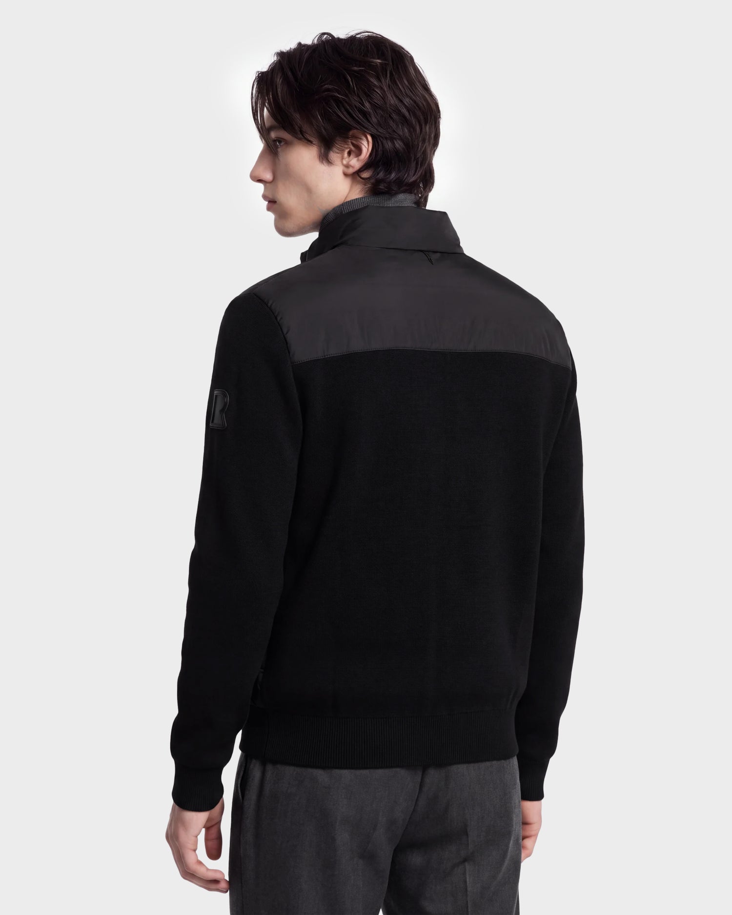 A young man with dark hair is seen from behind wearing the Rudsak EUGENE - 6124721 Black men's hybrid jacket with a high collar and gray pants, standing against a plain light background.
