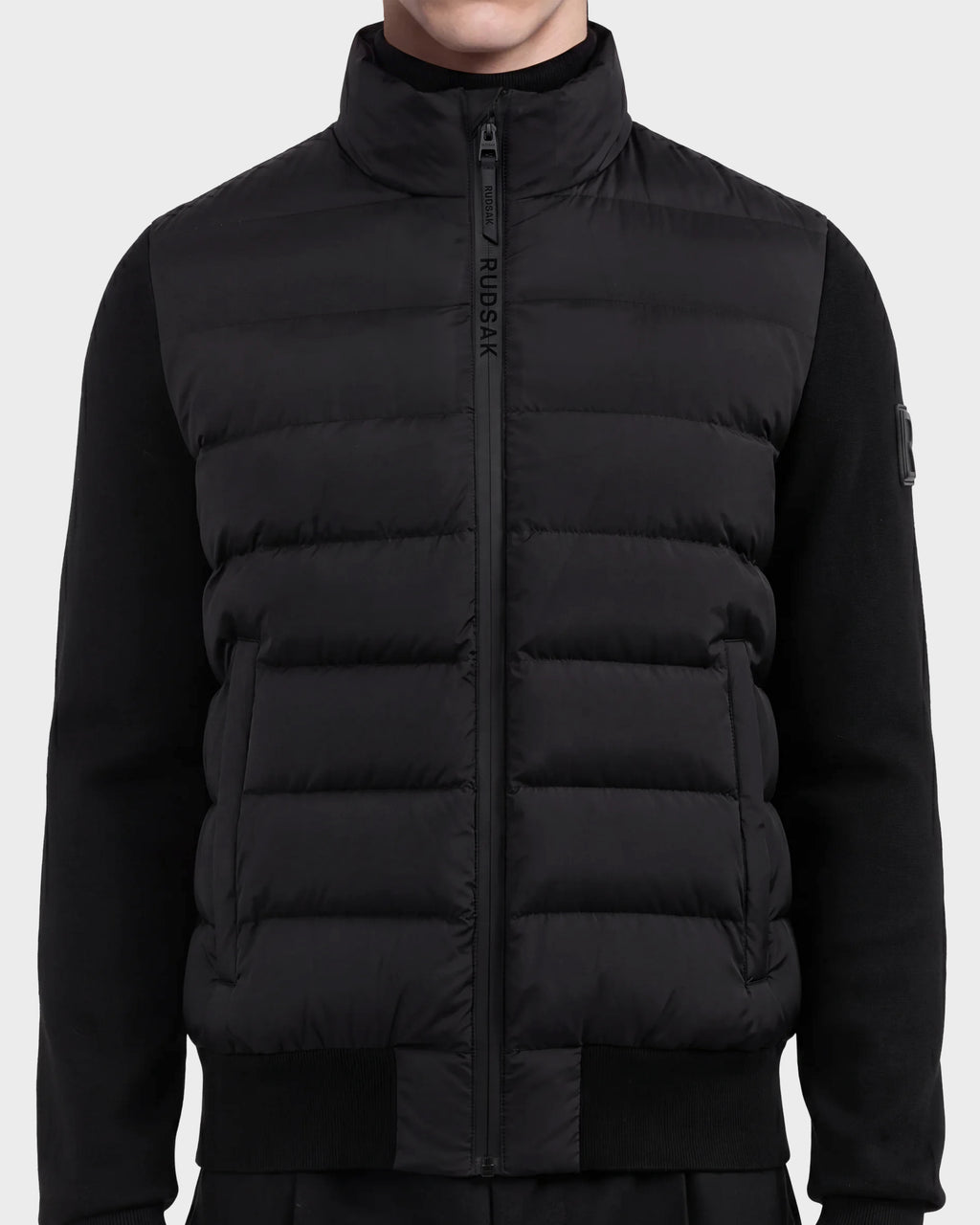 A person wearing the EUGENE - 6124721 Black men's hybrid jacket by Rudsak, featuring a high collar, zip closure with the Rudsak logo on the zipper flap, crafted from performance luxury fabric with ribbed cuffs and sides for a minimalist, modern look.