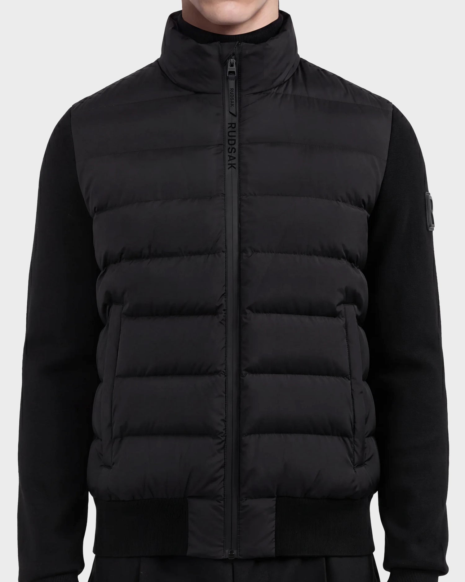 A person wearing the Rudsak EUGENE - 6124721 Black men's hybrid jacket, featuring a puffer-style design with a front zipper and high collar, over a long-sleeve black shirt; shown from the shoulders to the waist.