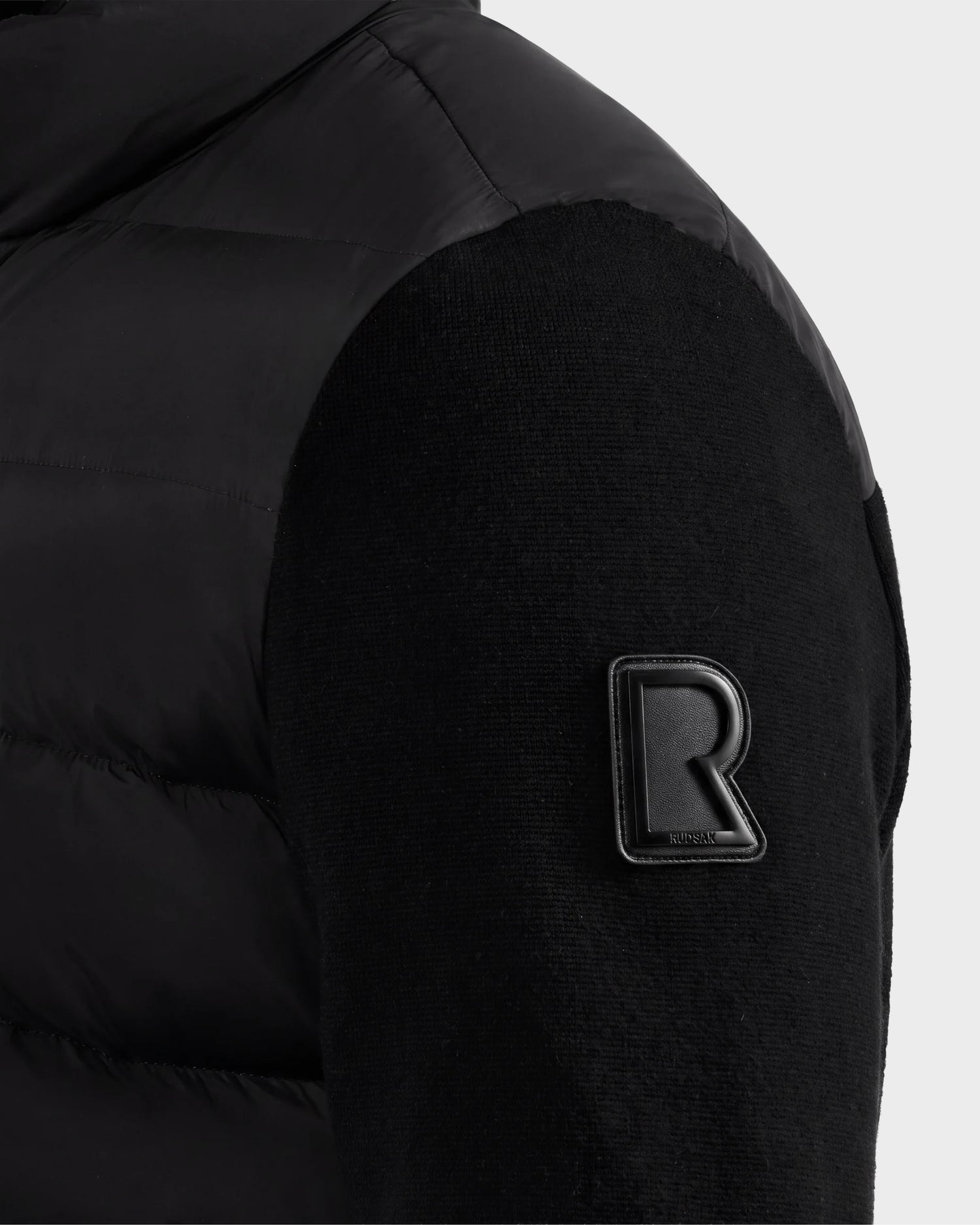 Close-up of a black padded sleeve from the EUGENE - 6124721 Black performance luxury jacket by Rudsak, featuring a raised black "R" patch near the upper arm with "Rubrik" engraved on its lower edge.