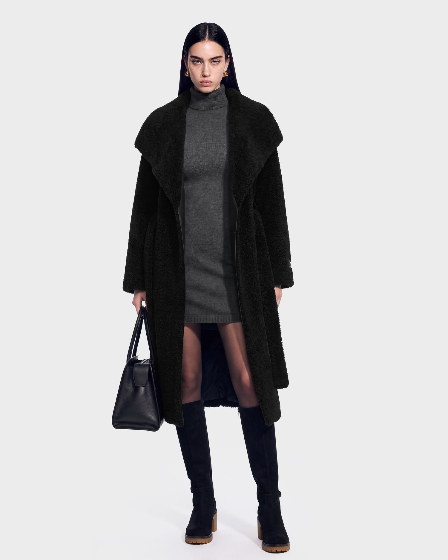 A woman wearing the FRANCIE - 8126873 Black long coat by Rudsak over a gray turtleneck dress, paired with black knee-high boots and a black handbag, stands against a plain light background with a neutral expression. This item is available for pre-order and is expected to ship by September 15th.