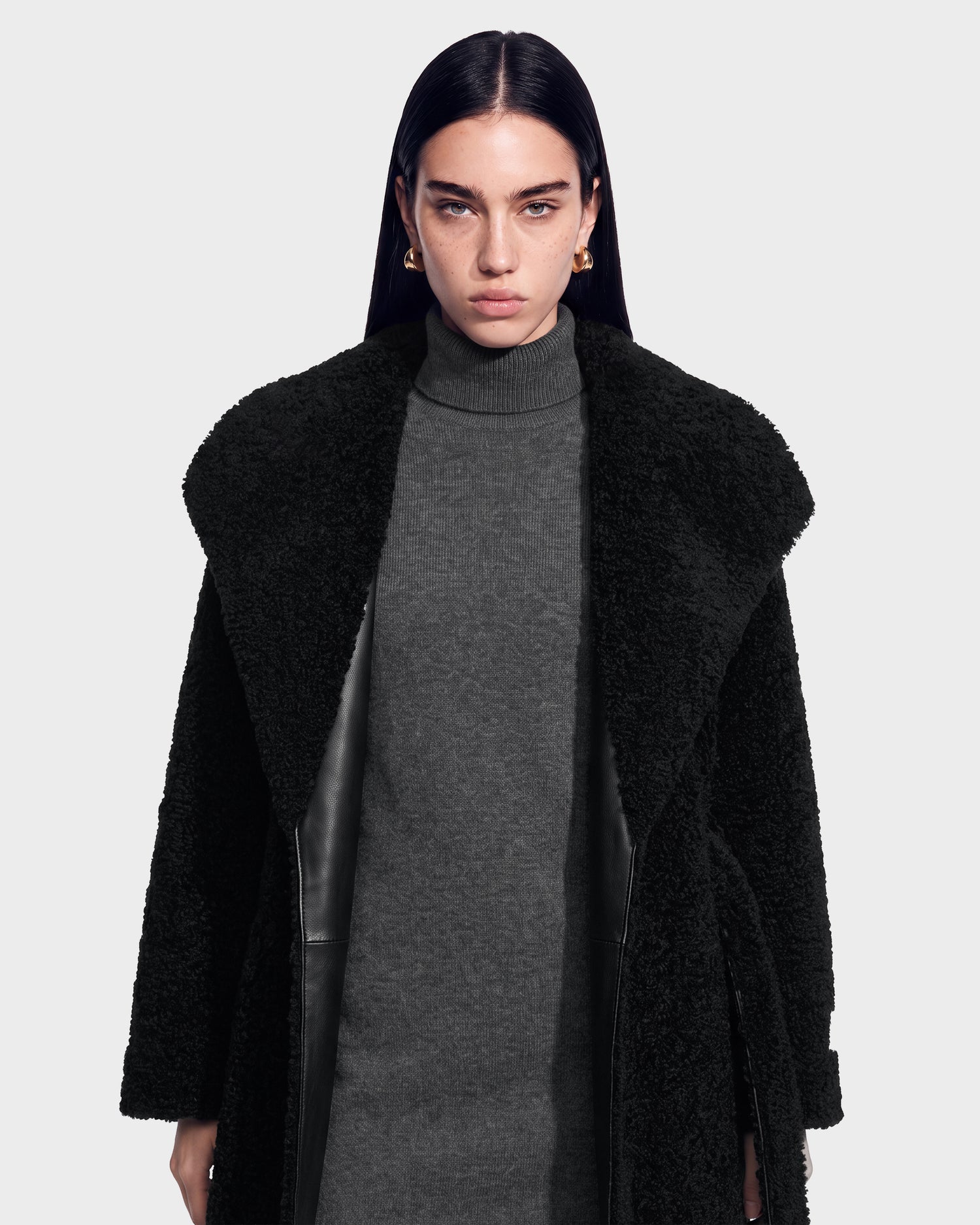 A woman with straight dark hair wears gold hoop earrings, a gray turtleneck dress, and the Rudsak FRANCIE - 8126873 Black textured coat, standing against a plain light background. This chic look featuring the FRANCIE coat is now available for pre-order and is expected to ship by September 15th.