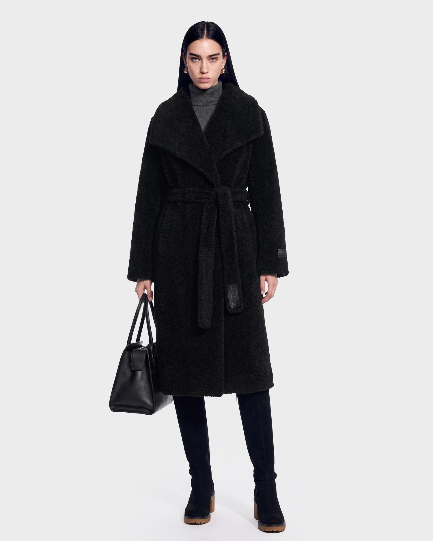 Wearing the FRANCIE - 8126873 Black belted coat by Rudsak, paired with black boots and a matching black handbag, this chic ensemble stands out against a simple light background. Now available for pre-order, the stylish look is expected to ship by September 15th.