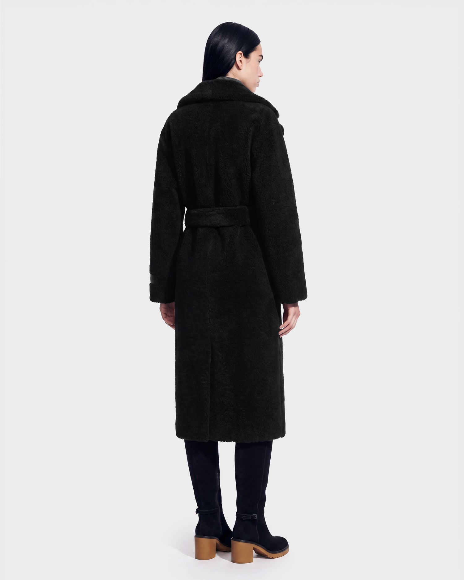 A person with long dark hair, dressed in Rudsak’s FRANCIE - 8126873 Black coat with a belt, black pants, and black boots stands facing away against a plain light background. This striking look is available for pre-order and is expected to ship by September 15th.