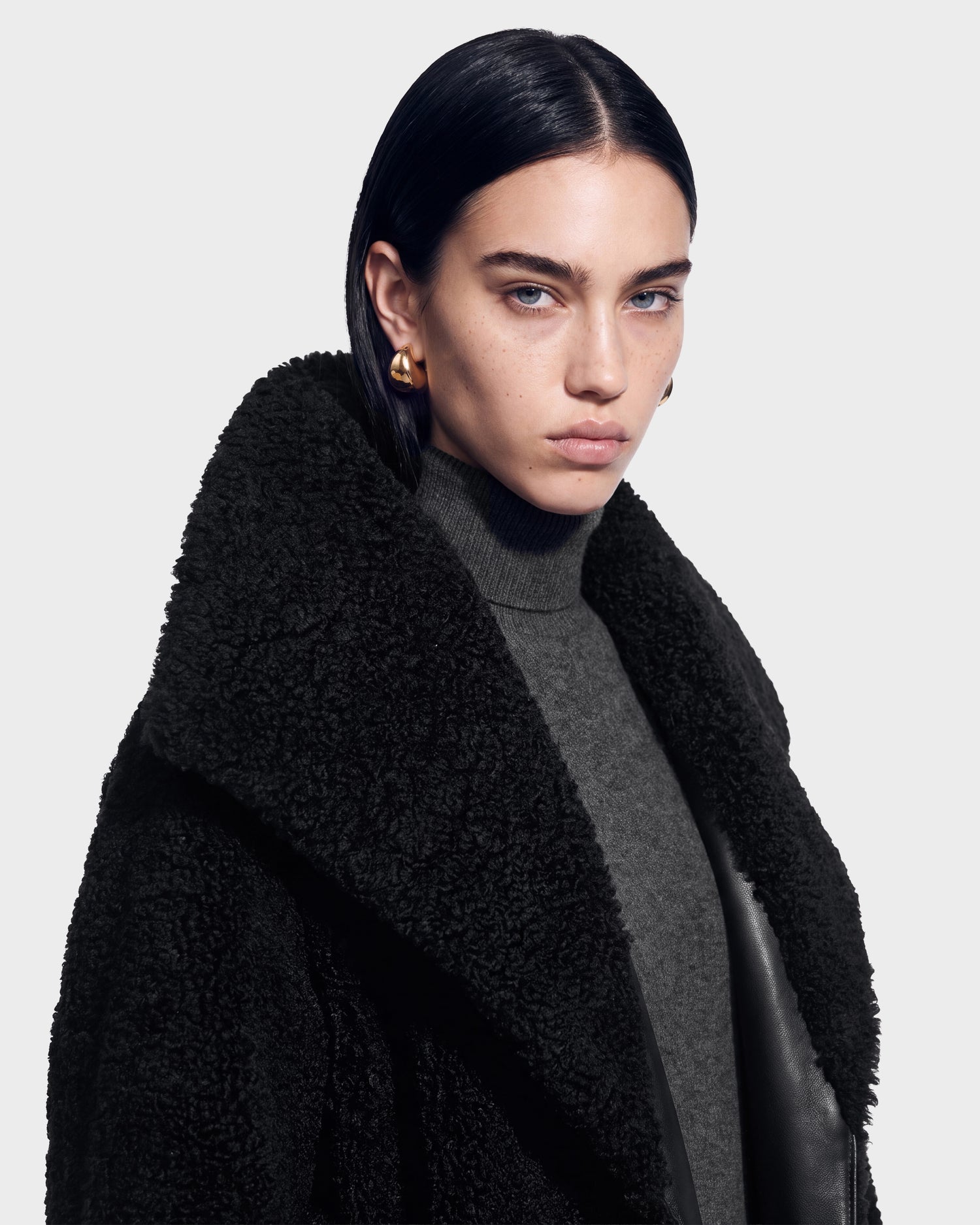 A person with straight dark hair, gold hoop earrings, and a serious expression wears the textured black FRANCIE - 8126873 coat by Rudsak over a gray turtleneck sweater, posing against a plain light background—a look now available for pre-order and expected to ship by September 15th.