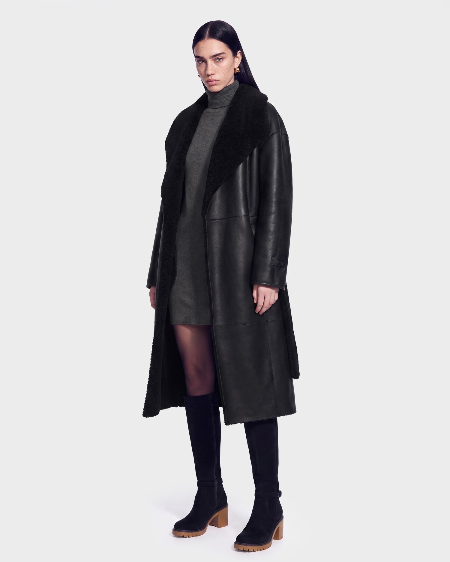 A woman stands facing forward, wearing the Rudsak FRANCIE - 8126873 Black long coat with a wide collar over a dark gray dress, black tights, and black heeled boots. She has long dark hair and a neutral expression against a plain background. The FRANCIE coat by Rudsak is now available for pre-order and is expected to ship by September 15th.