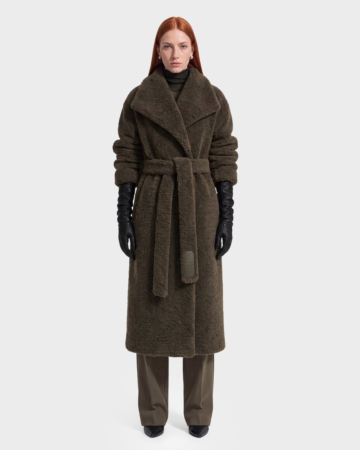 A woman with long red hair wears the Rudsak FRANCIE - 8126873 Olive belted, textured vegan leather coat over dark clothing and black gloves, standing against a plain white background.