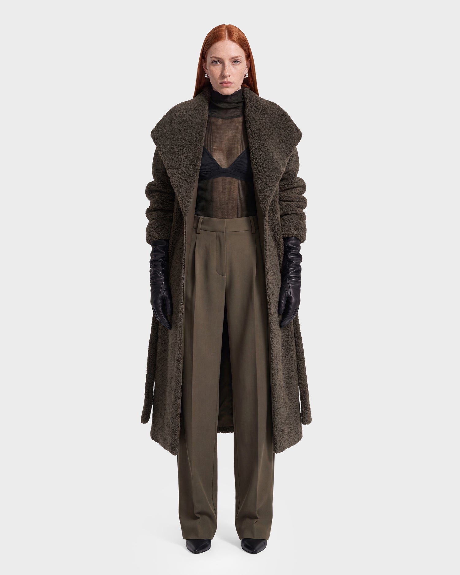 A person with long red hair stands against a plain white background wearing the Rudsak FRANCIE - 8126873 Olive wide-leg trousers, paired with a brown faux shearling textured coat, sheer black top layered over a black bra, black gloves, and black shoes.