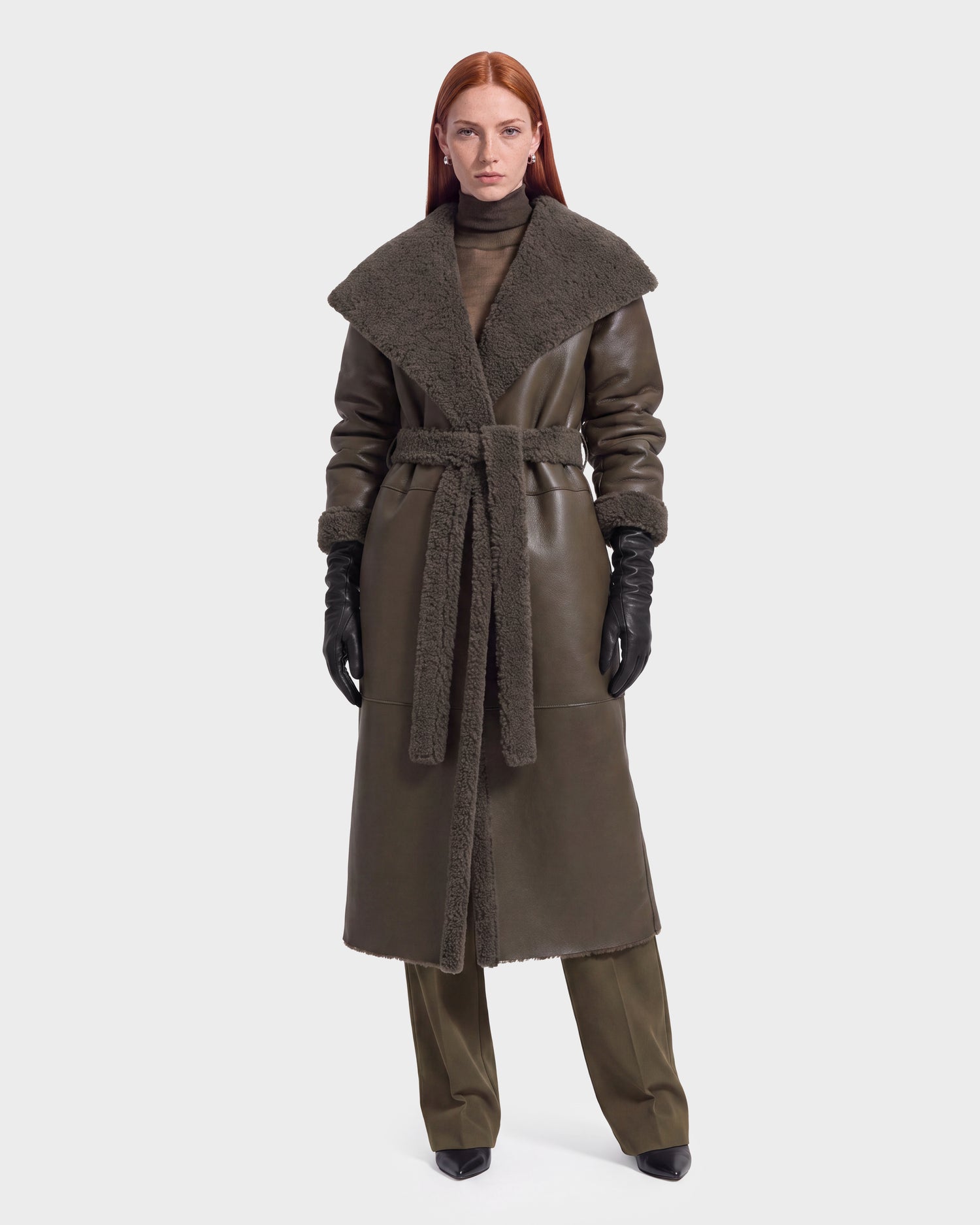 A person with long red hair wears the Rudsak FRANCIE - 8126873 Olive long dark green faux shearling coat with a wide collar, matching belt, gloves, and pants, standing against a plain white background.