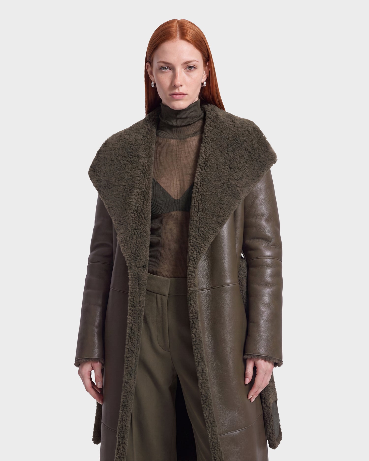 A woman with long red hair wears the Rudsak FRANCIE - 8126873 Olive faux shearling coat over a sheer turtleneck and pants, standing against a plain light background.
