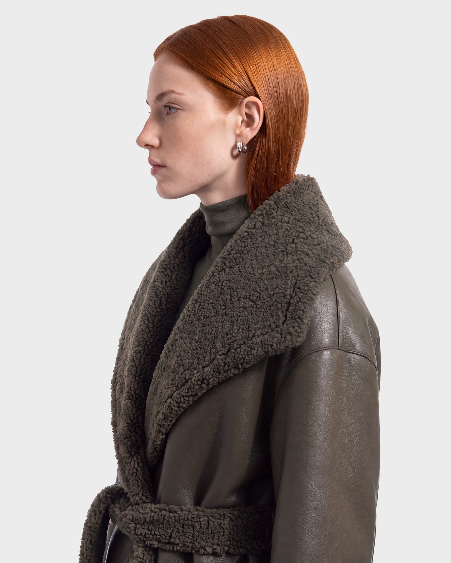 A woman with straight red hair, facing left against a plain light background, wears the Rudsak FRANCIE - 8126873 Olive faux shearling coat over a turtleneck. She accessorizes with small hoop earrings and has a neutral expression.