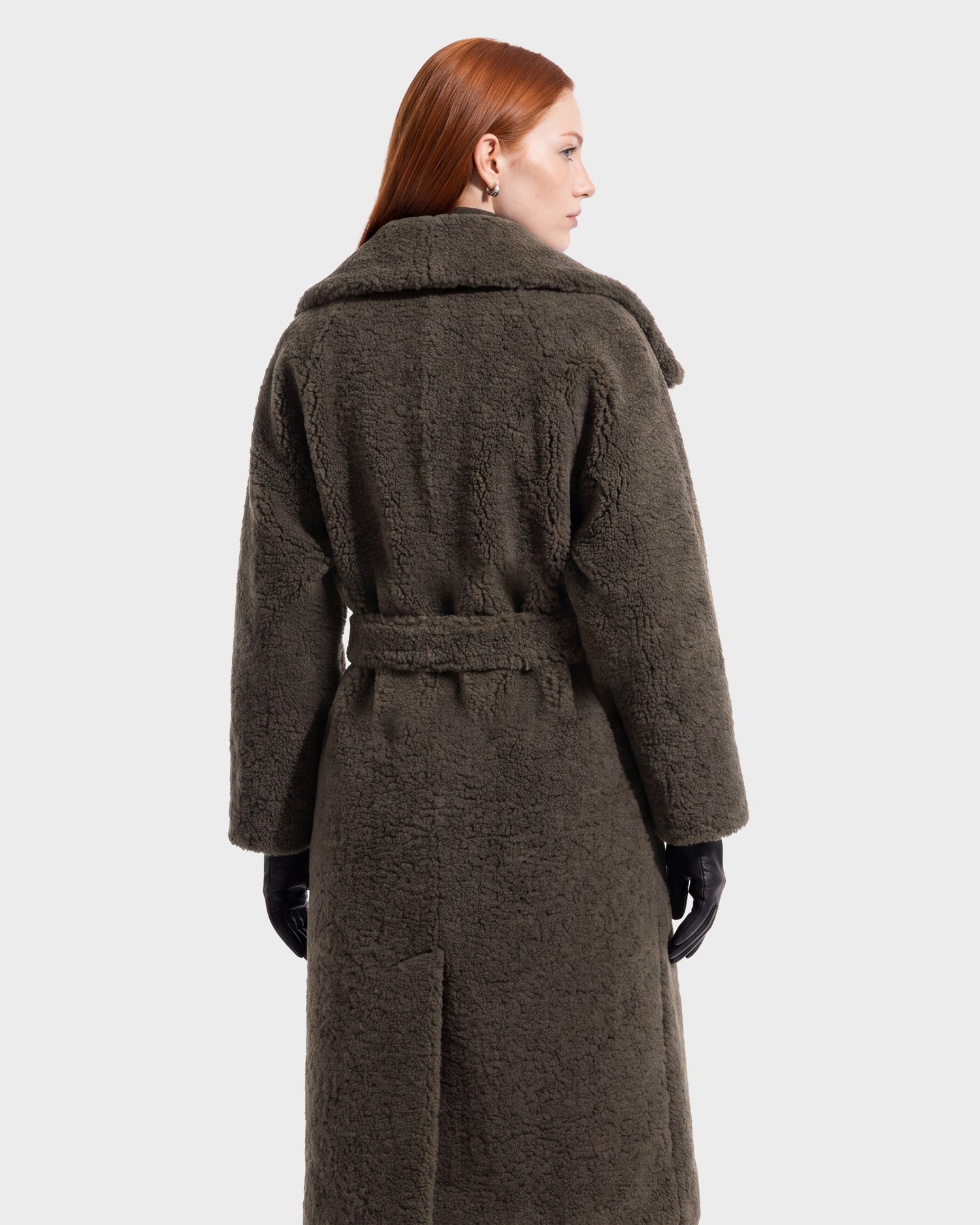 A person with long red hair, wearing the Rudsak FRANCIE - 8126873 Olive vegan leather long coat and black gloves, is shown from the back against a plain white background.