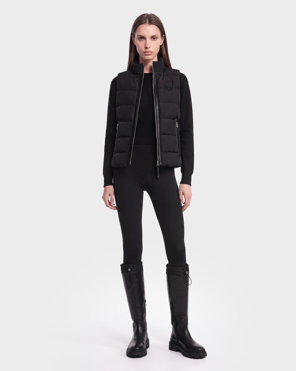 A person stands facing forward against a plain white background, wearing the GABRIELA HV - H125507 Black puffer vest by Rudsak over a black long-sleeve top, paired with black pants and knee-high black boots.