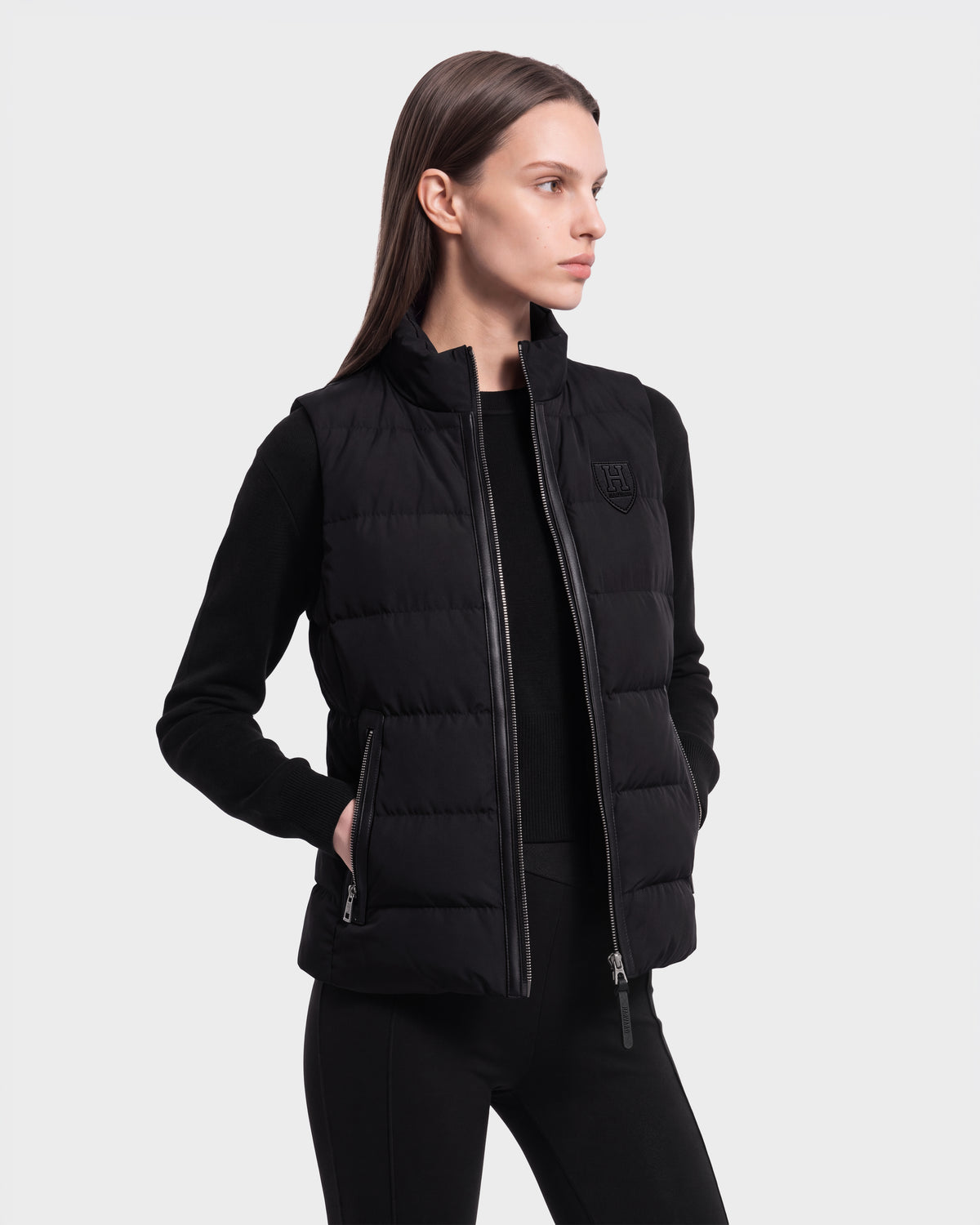 A woman with straight brown hair models the Rudsak GABRIELA HV - H125507 Black RDS-certified down puffer vest, layered over a black long-sleeve shirt and black pants. She stands with one hand in her pocket against a plain light background.