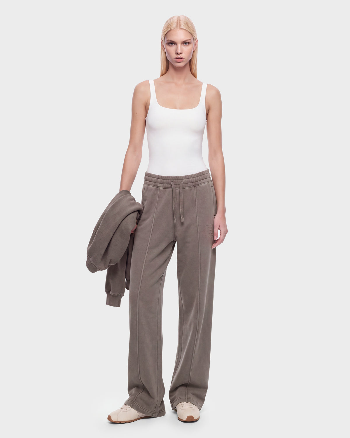 A woman with long blonde hair wears a white tank top, loose GAELLE HV - H526902 Taupe jogger sweatpants by Rudsak, and beige sneakers. She holds a matching taupe sweatshirt in her left hand and stands against a plain white background.