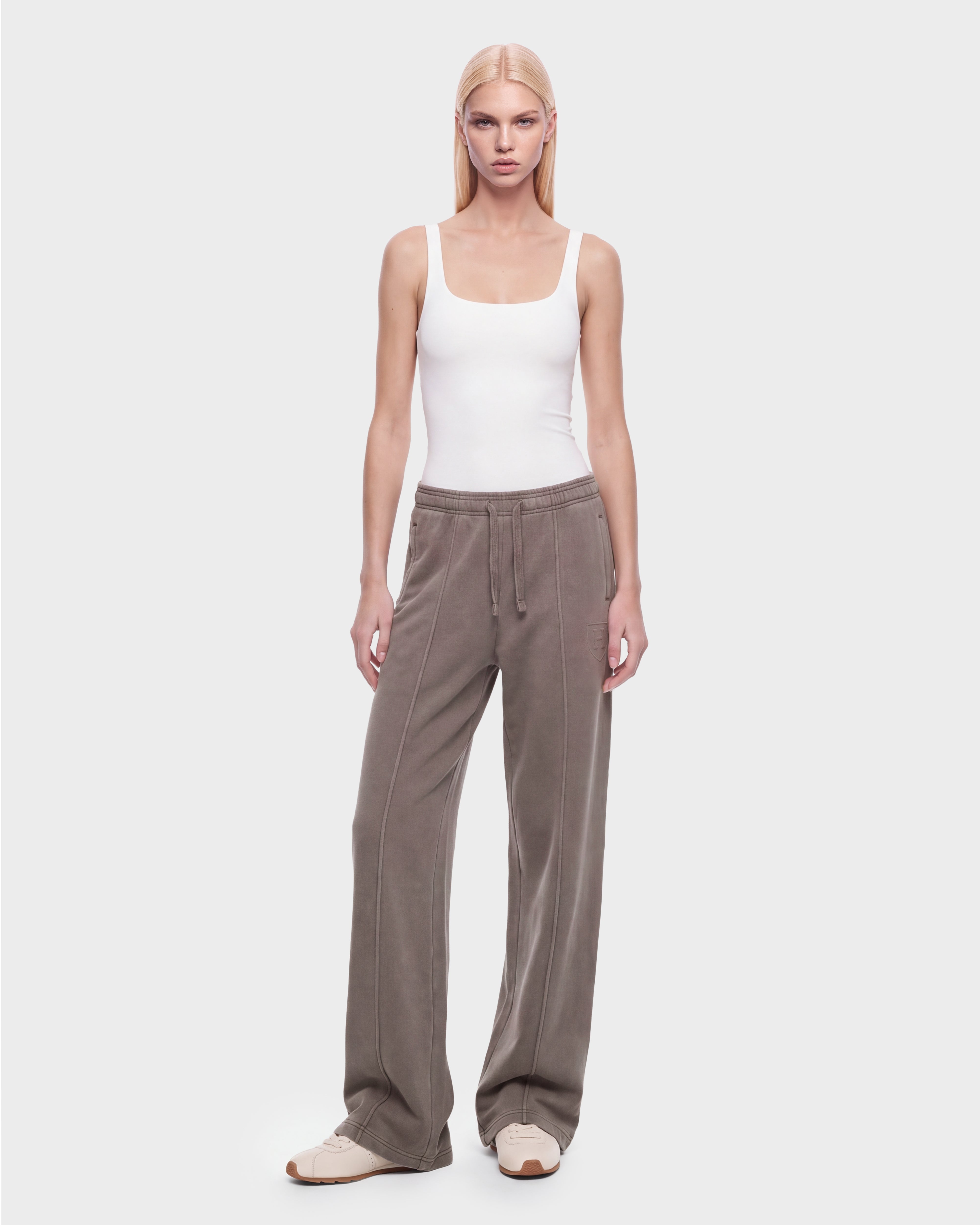 A person with straight blonde hair wears a white tank top, Rudsak GAELLE HV - H526902 Taupe wide-leg pants featuring visible seams reminiscent of heavyweight jersey joggers, and beige sneakers, standing against a plain light gray background.