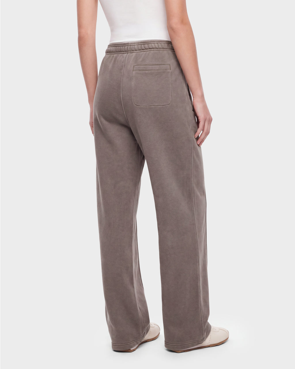 A person is shown from the back against a plain background wearing the Rudsak GAELLE HV - H526902 Taupe pants—loose, taupe wide-leg trousers with an elastic waistband and a single back pocket, reminiscent of heavyweight jersey joggers—paired with a white sleeveless top and white shoes.