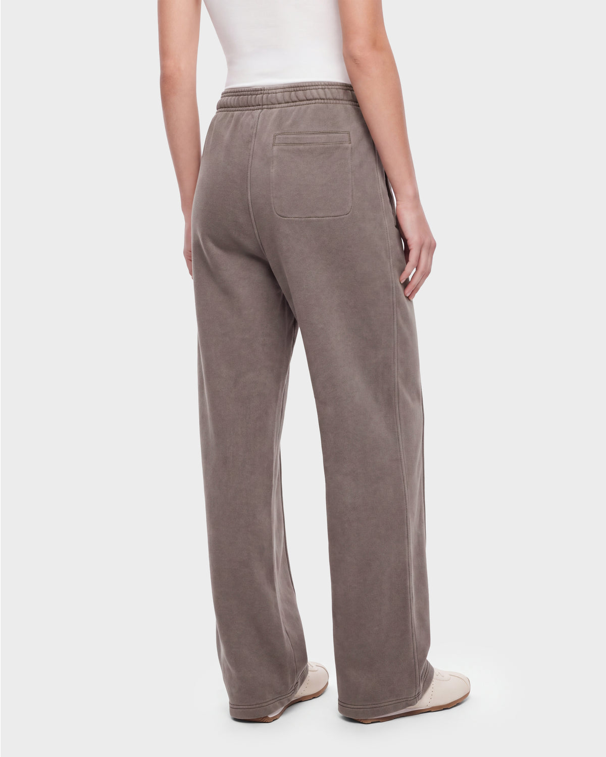 A person is shown from the back against a plain background wearing the Rudsak GAELLE HV - H526902 Taupe pants—loose, taupe wide-leg trousers with an elastic waistband and a single back pocket, reminiscent of heavyweight jersey joggers—paired with a white sleeveless top and white shoes.